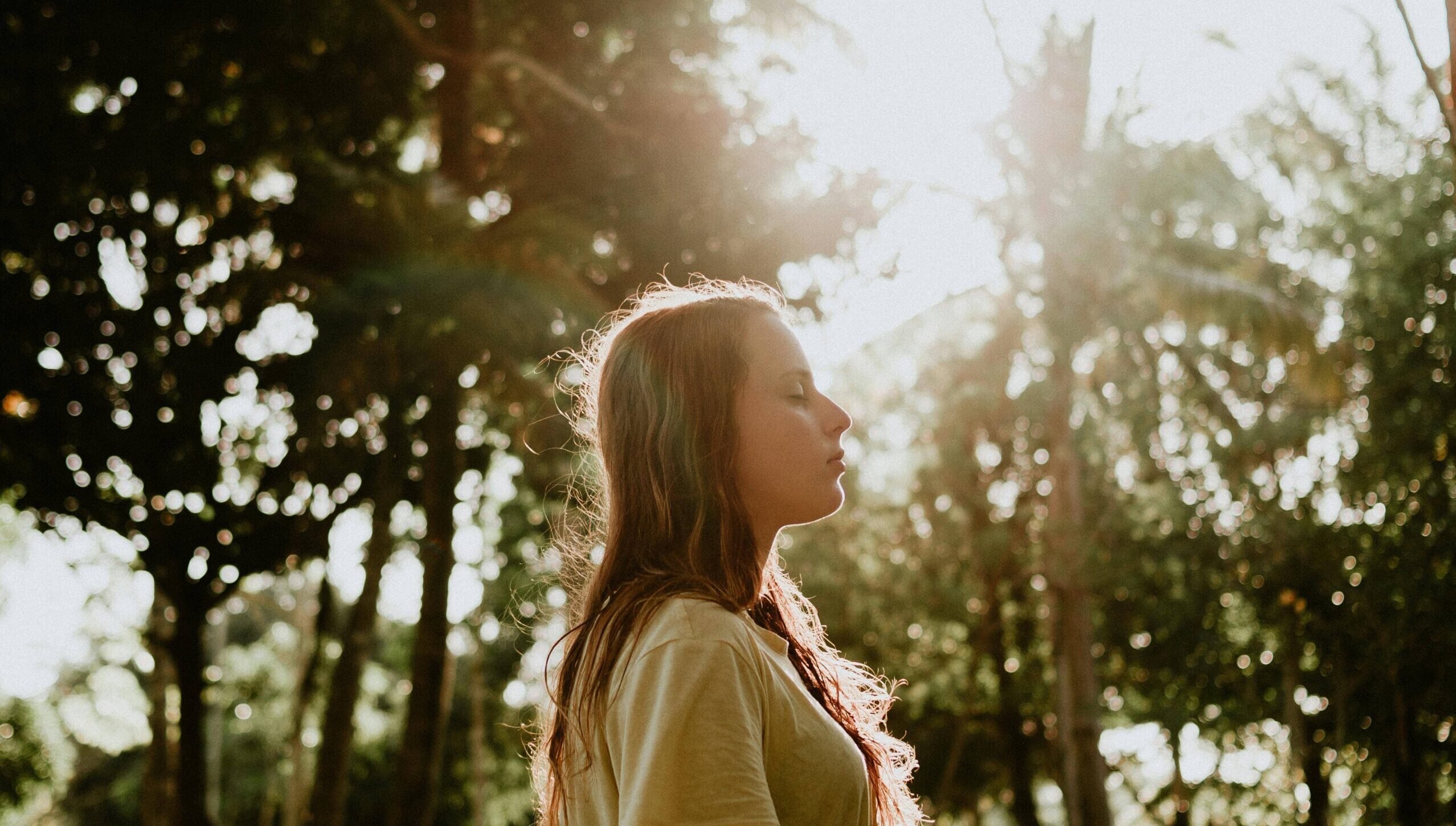 Woman standing in forest with eyes closed and sun beaming down her.